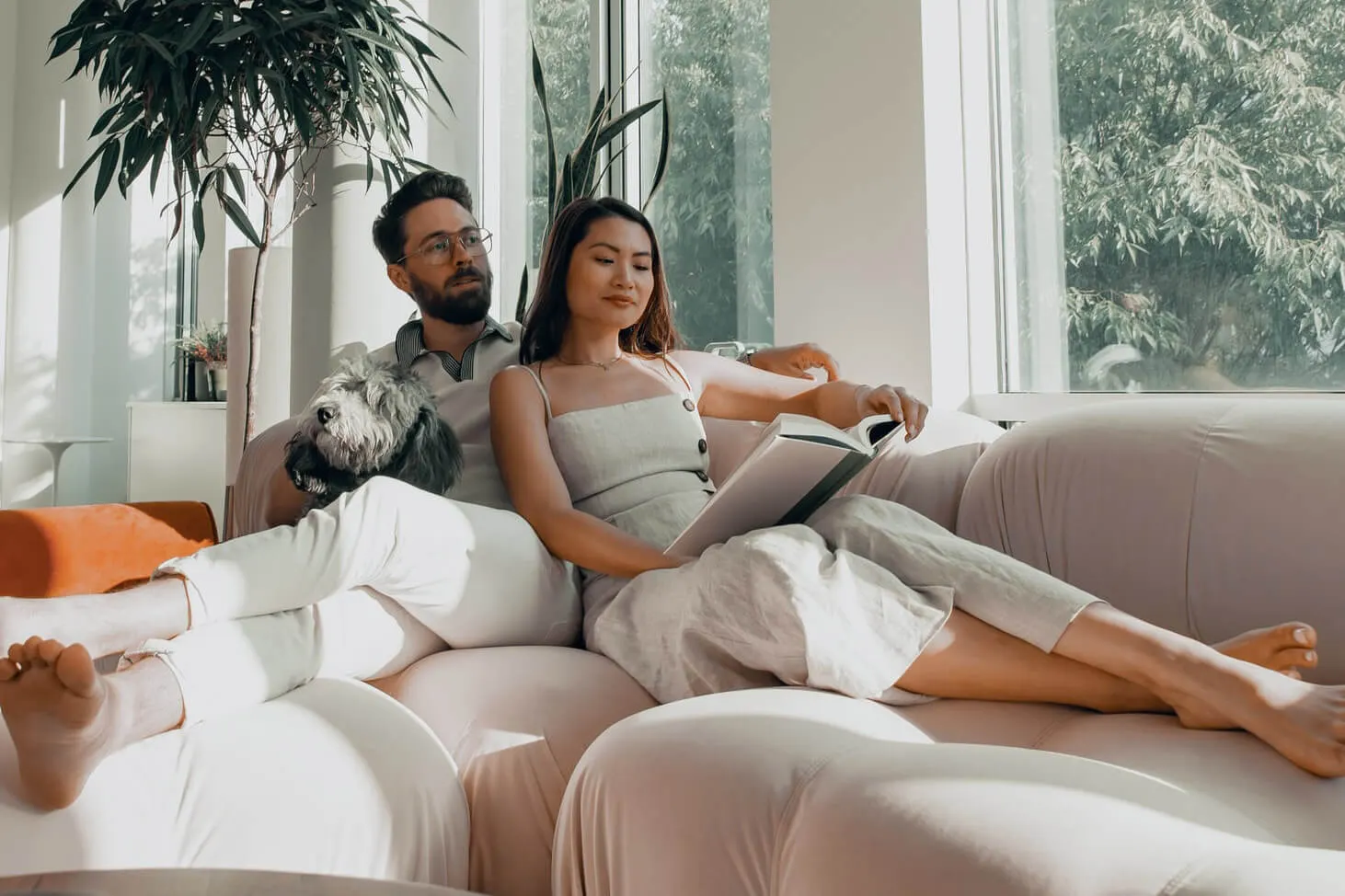 A couple relaxing on a comfortable gray sofa in a sunlit living room. They are leaning against each other, sharing a warm drink, and looking relaxed with soft smiles.
